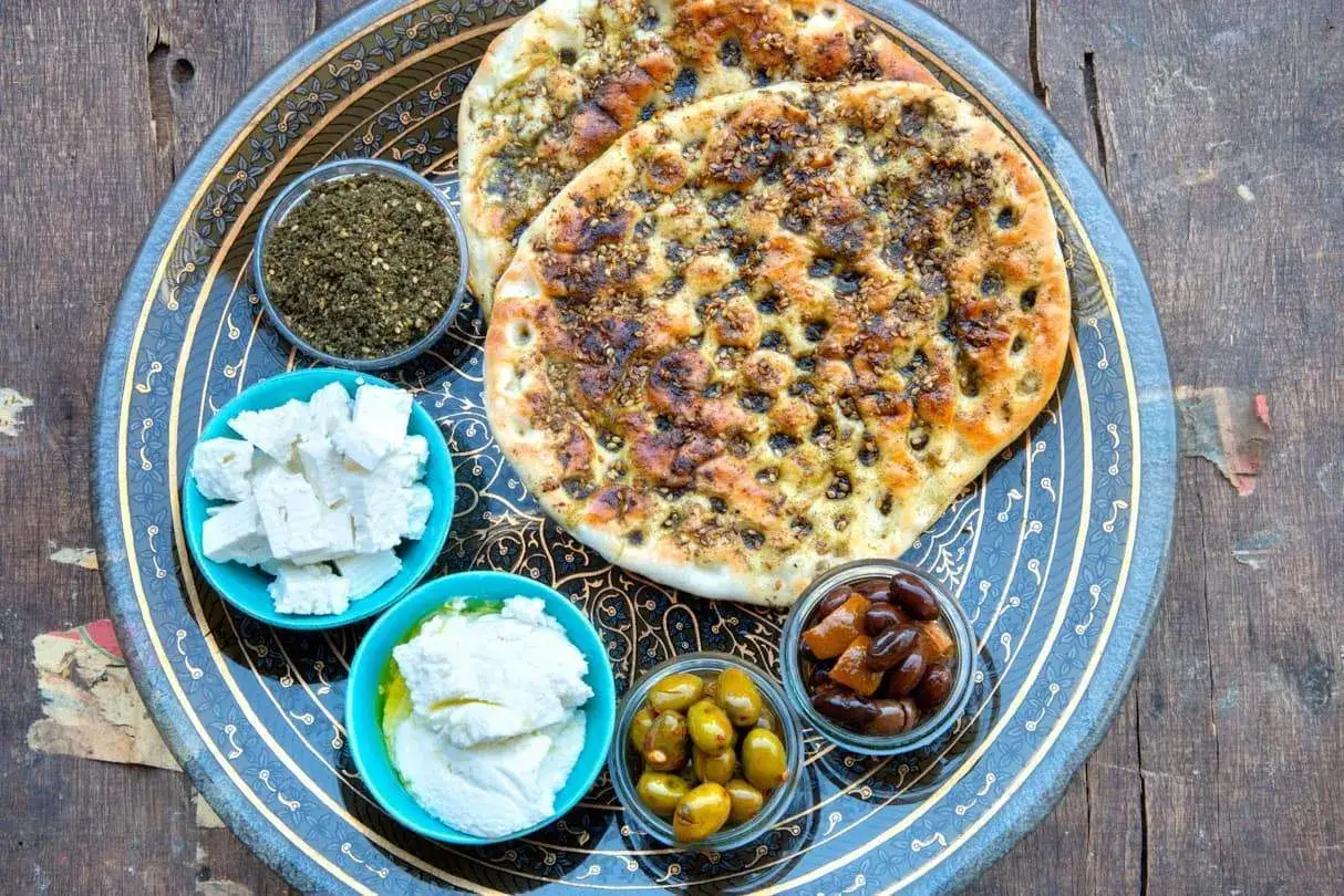 Fladenbrot mit Zatar, Oliven, Feta und Frischkäse auf einem verzierten Tablett. Was ist Zatar? Eine würzige Kräutermischung.
