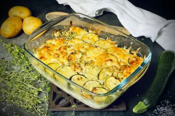 Goldbrauner Zucchini-Kartoffel-Auflauf mit Käse überbacken, garniert mit frischem Thymian. Daneben liegen Kartoffeln und eine Zucchini.