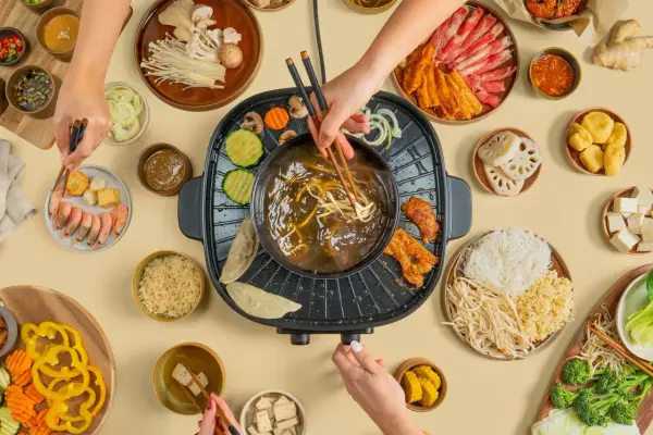 Hände greifen nach Zutaten für einen Hot Pot. Fleisch, Gemüse, Garnelen und Nudeln werden in die köchelnde Brühe getaucht.