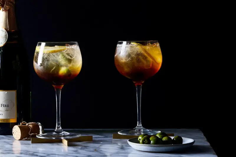 Zwei erfrischende Cynar Spritz Cocktails mit Eis, Zitrone und Oliven, daneben eine Champagnerflasche.