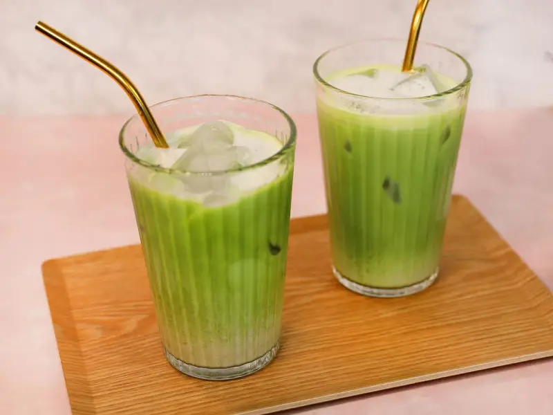 Zwei erfrischende Iced Matcha Latte mit goldenen Strohhalmen auf einem Holztablett. Perfekt für ein schnelles iced matcha latte rezept.