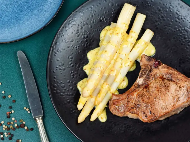 Ein saftiges Schweinekotelett liegt neben einem Haufen cremigen Spargel in der Pfanne, beträufelt mit einer würzigen Sauce.
